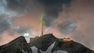 A telecom tower stands atop the Säntis mountain in Switzerland against a cloudy sky. A green laser marks the path of the powerful laser in this story.