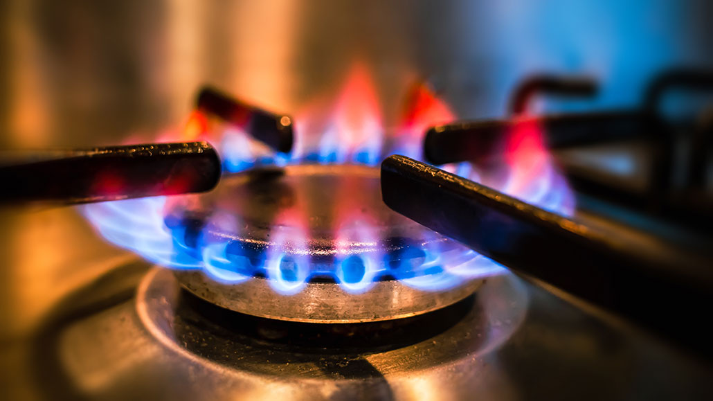 A close-up photo of a gas flame on a stove top burner. The flame is blue at the bottom and red at the tip of the flame.