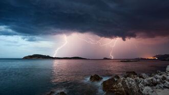 A violent thunderstorm approaching over the Adriatic Sea, with the lights of Dubrovnik Old Town visible in the distance.