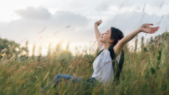 a photo of a young woman with pale skin and long dark hair sitting in a windy field and lifting her arms to the sky