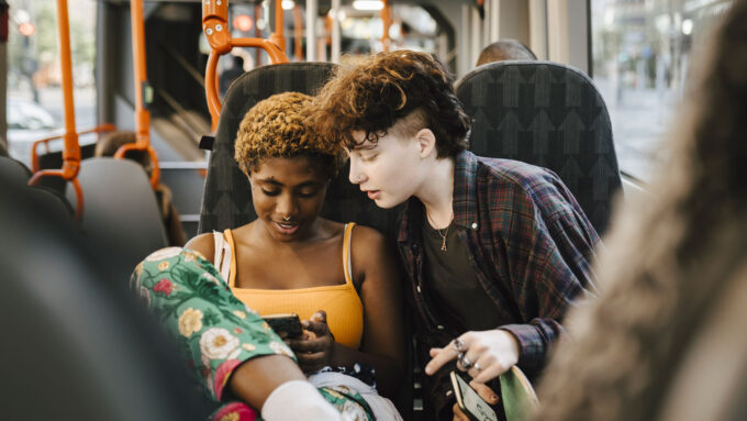 two teens sit side by side on a bus, one leaned over the other's shoulder to look at their phone together