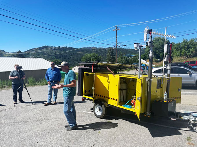 a photo of a yellow vehicle on wheels with satellite antennas poking out of the back. A man in a green shirt and cap is talking to an audience off screen.