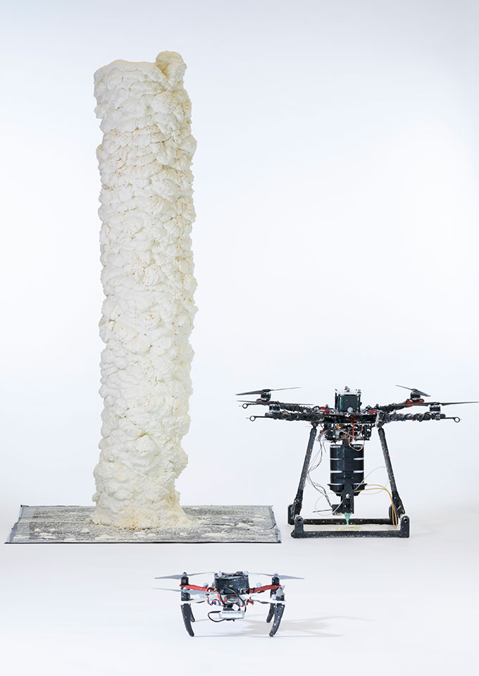a tower of cylindrical white foam with the drone that printed it sitting next to it. In the foreground is Scan-drone, a smaller drone.