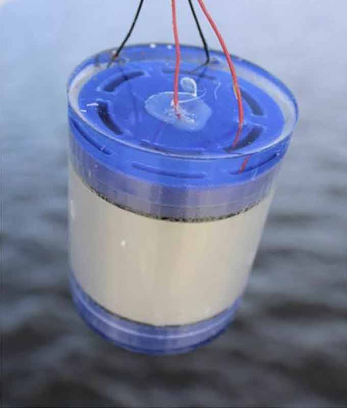 a photo of an underwater transmitter, a white and blue cylinder hanging over water, wires extend from the top of the cylinder