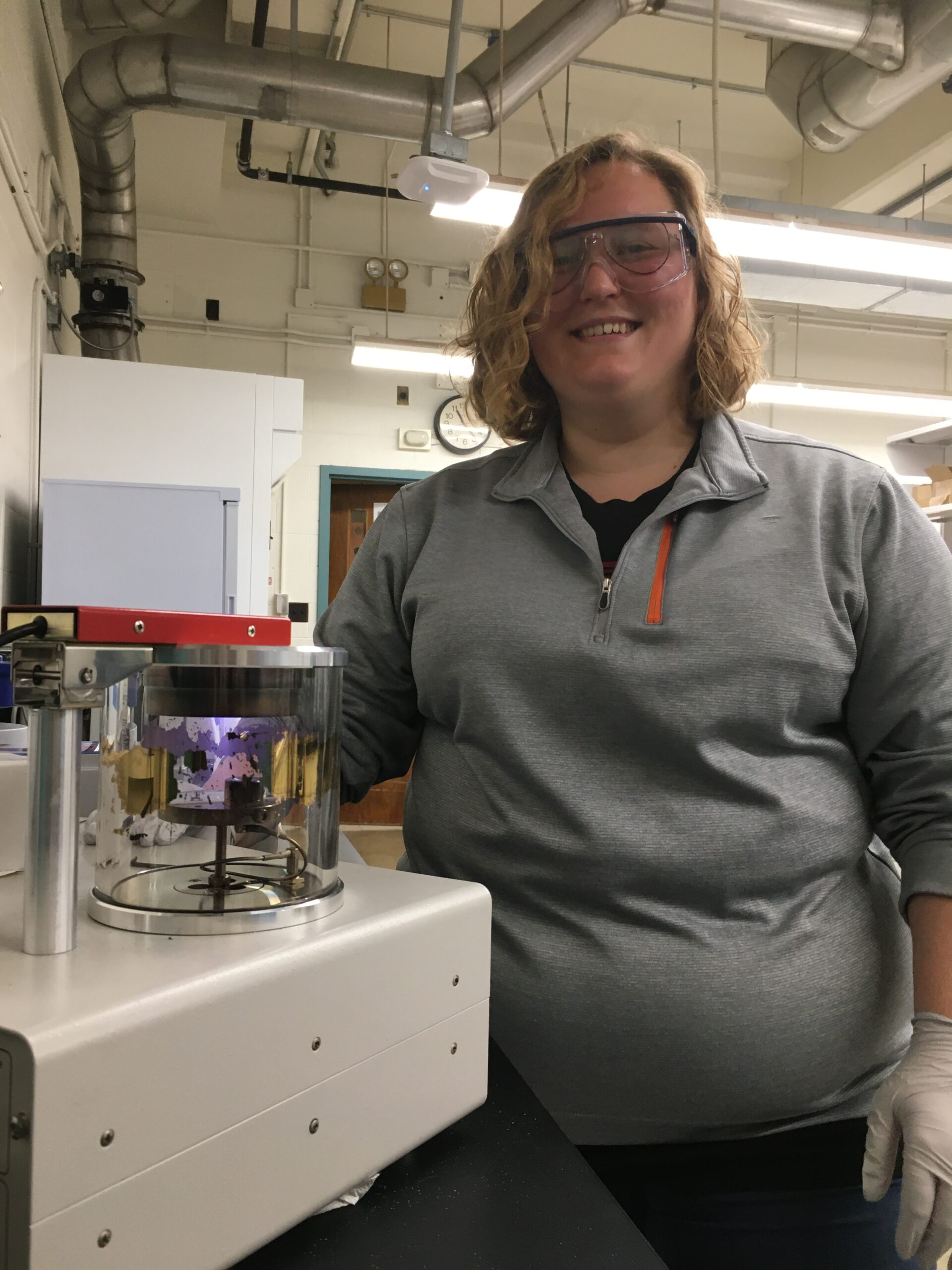 a young blonde woman wearing goggles stands by a piece of machinery in a lab