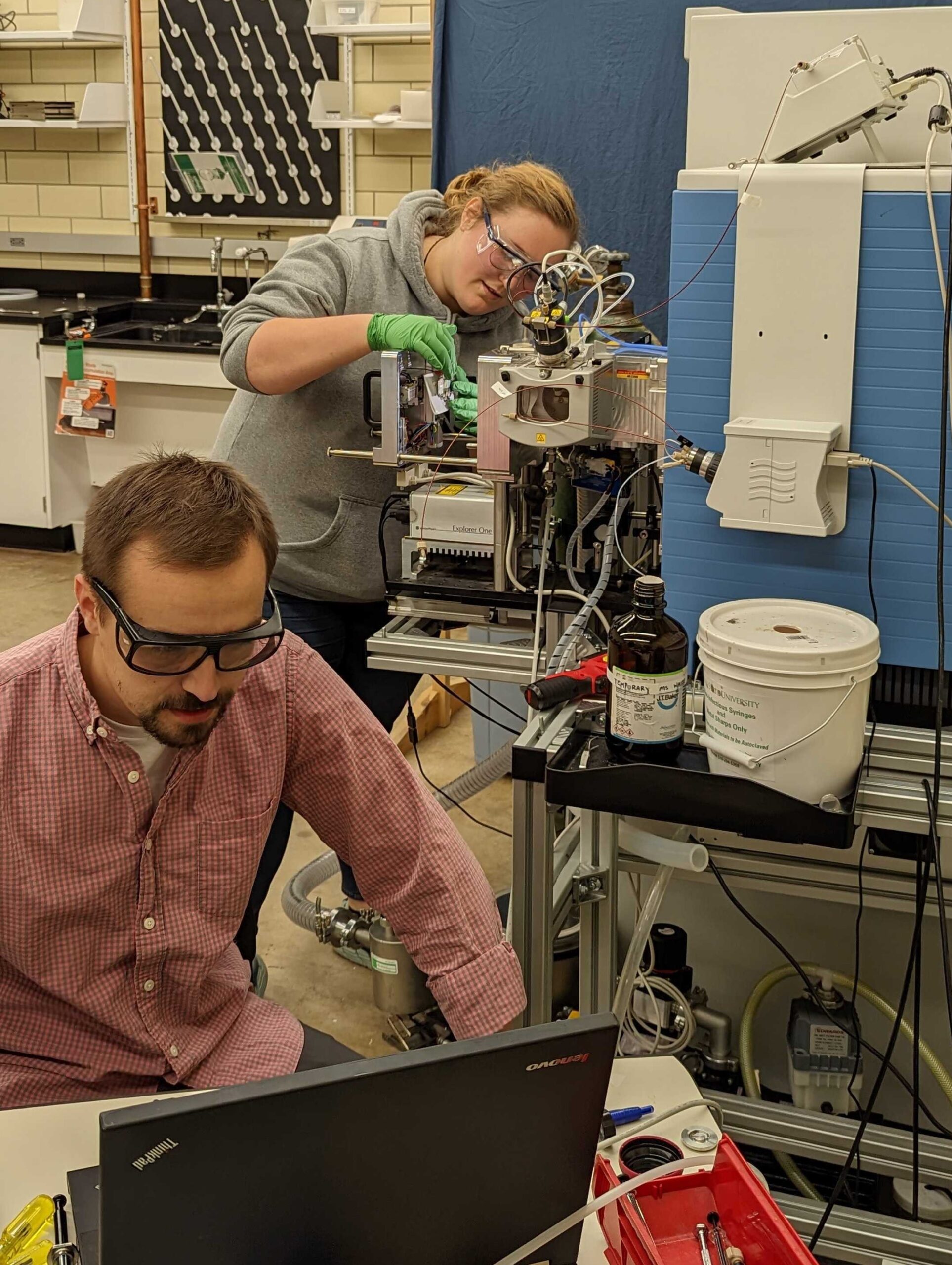 in a lab cluttered with equipment, a young man and woman wearing goggles look at a computer and work on machinery