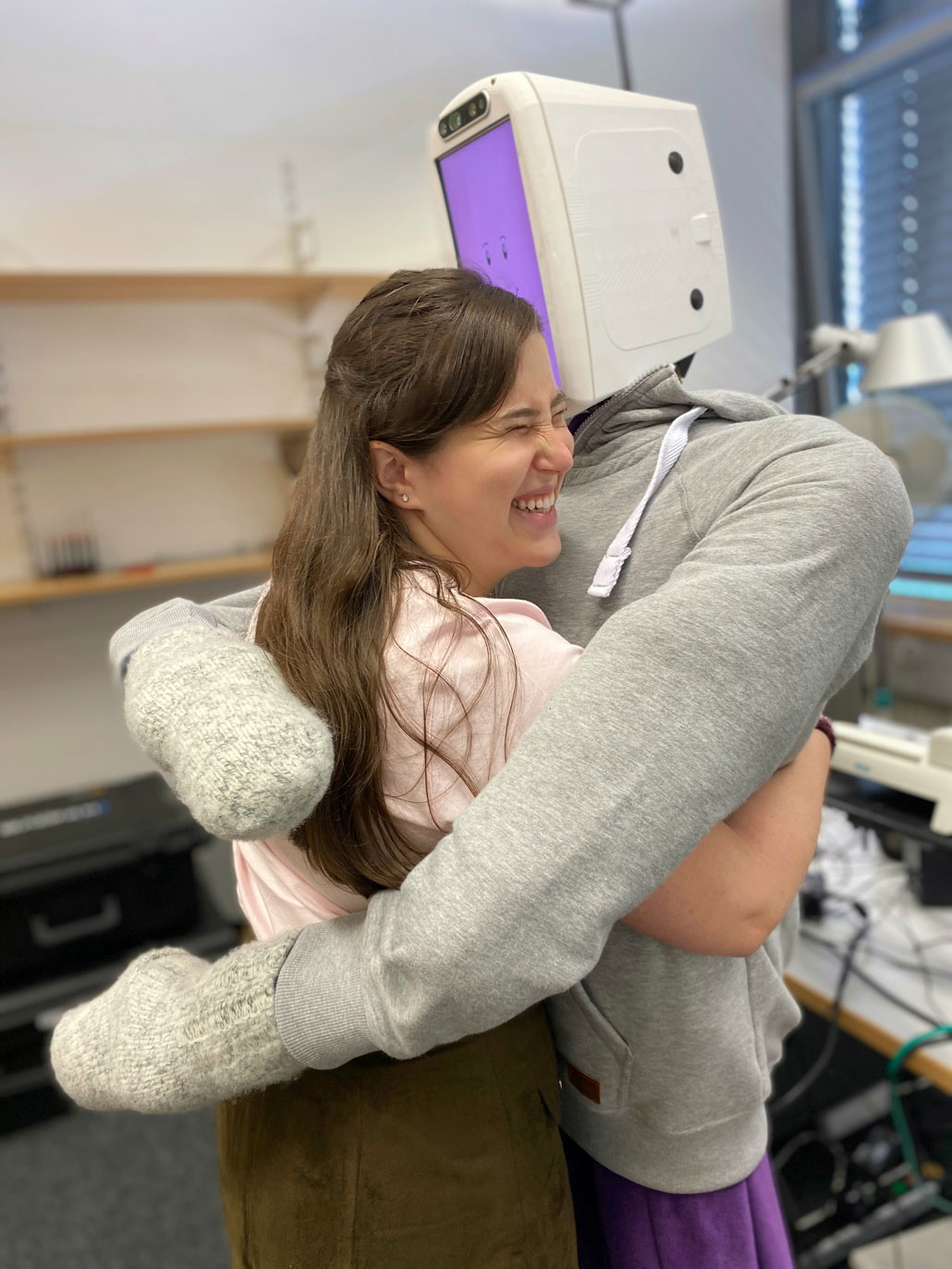 A white woman with long brown hair is smiling and hugging a soft-armed robot. The robot's screen-head is lit up purple with a smiling emoji face and it is hugging her back.