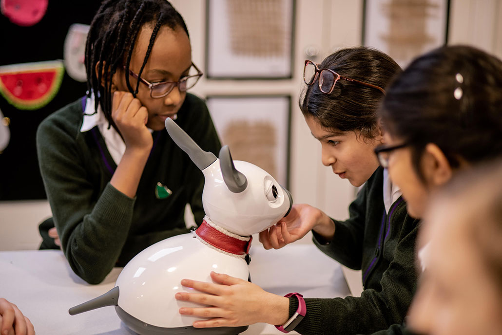 a group of students sits at a table playing with a small animal shaped robot with long ears 