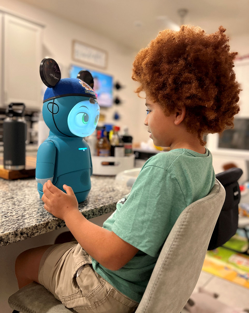 Moxie, a small blue robot wearing MIckey Mouse ears stands on a countertop and interacts with Rocco, a brown-skinned boy with curly red-brown hair in an afro