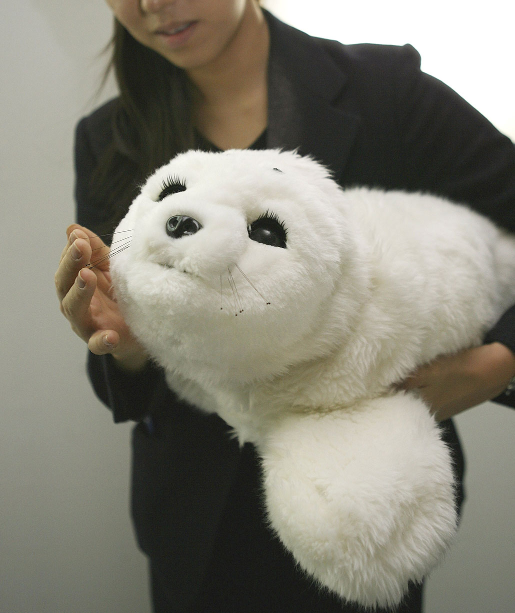 Paro, an ultra-adorable soft robot seal, is being held by a young Japanese woman in a suit. She is about to stroke its cheek, and its face is turned towards the viewer.