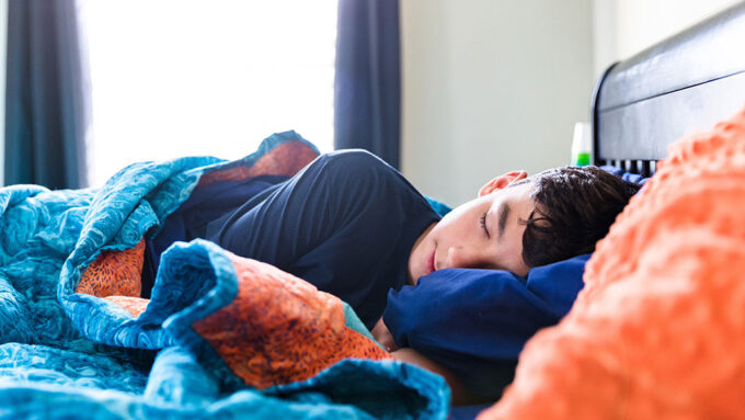 a photo of a teenager laying in bed asleep. They are snuggled under a cozy blue and orange fleece blanket