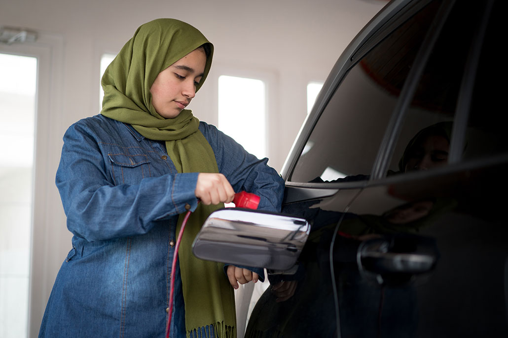a girl in a blue jean tunic wearing a green headscarf is plugging in her electric car for charging