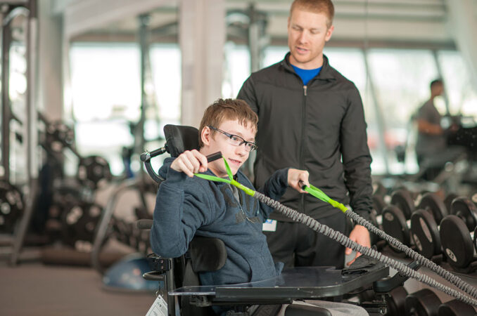 Boy in wheelchair pulls on resistance bands as his personal trainer supervises