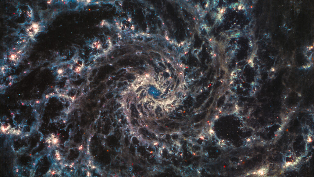An image from the James Webb Space Telecope showing face-on spiral galaxy, NGC 628, with its whorls of colored gas and dust, pockmarked with dark areas.