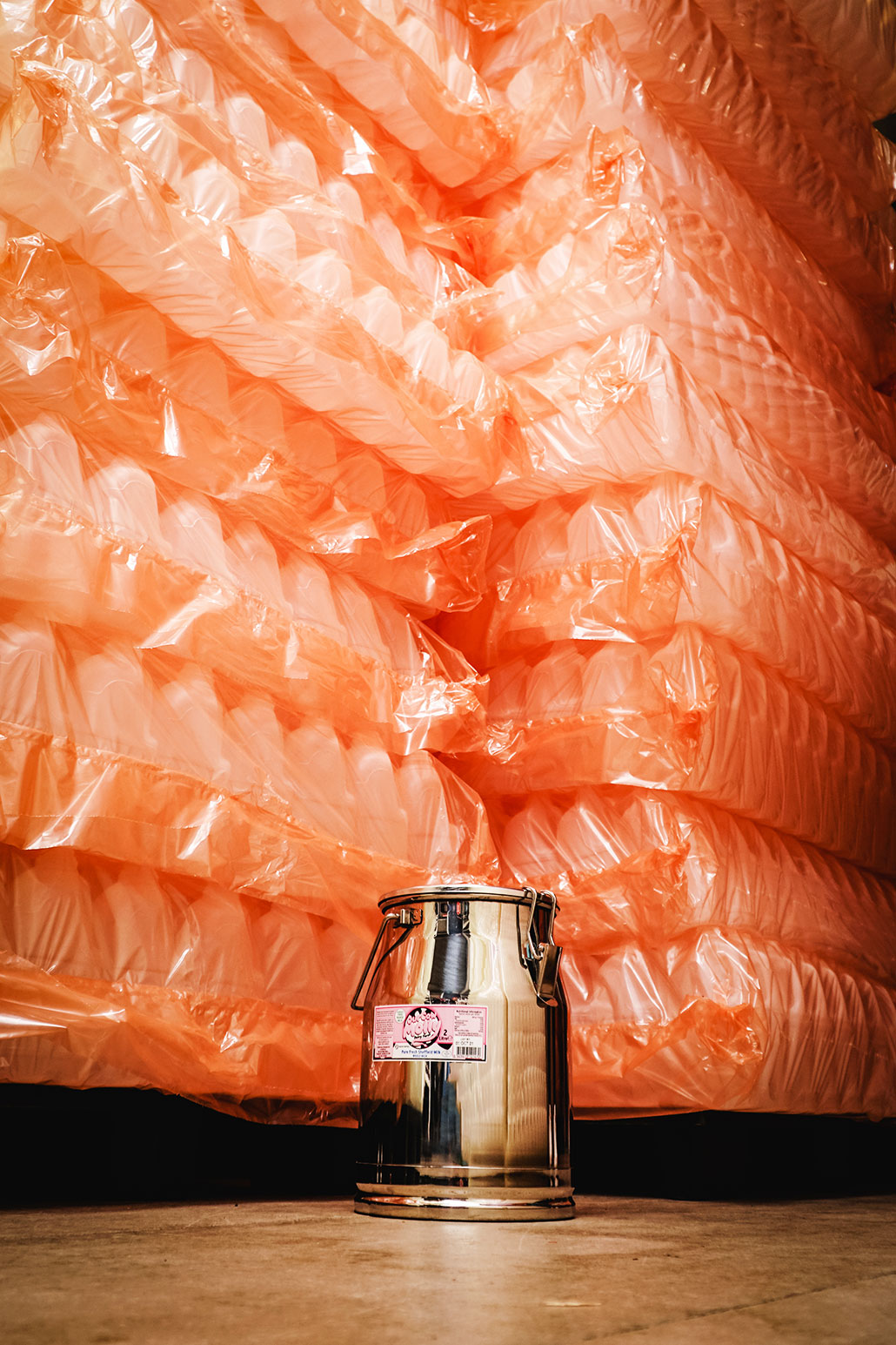 a silver metal milk jug on a warehouse floor in front of stacks and stacks of plastic milk jugs in orange plastic