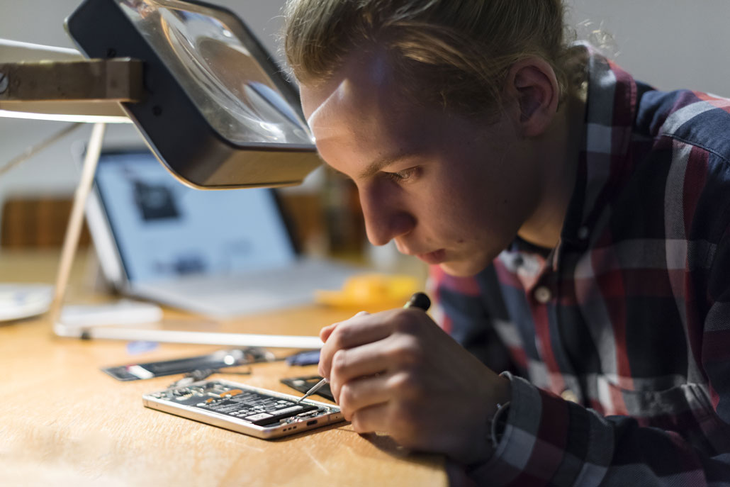 A photo of a young man bent over a phone that has been opened up and taken apart. A laptop is in the background. Various tools are scattered on the table. Above his head is a magnifier.