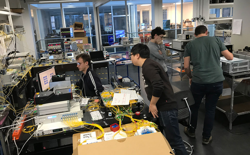 a photo of several adult males clustered in a lab working together. Various technology, wires and cables are on tables all around them