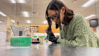 a high-school girl wearing a green sweater and glasses uses a pipette to deposit a liquid on a small paper sensor on a classroom desk