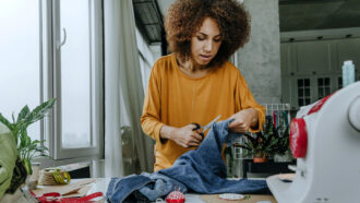 a photo of a young woman with an afro standing at a sewing table. She is looking down at her hands as she cuts a pair of jeans into useable fabric. Various sewing tools litter the table. A sewing machine is to the right of the picture.