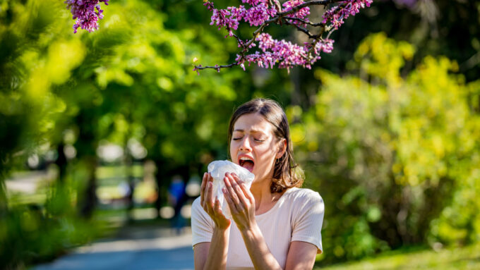 A woman surrounded by greenery and flowering trees prepares to sneeze into a tissue