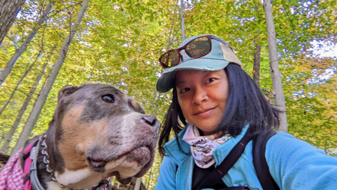 a woman wearing a baseball cap, jacket and sunglasses on her head takes a selfie in a forest with a dog