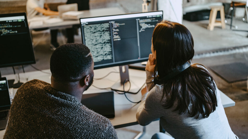 a teen boy and teen girl look at computer code written on a desktop screen