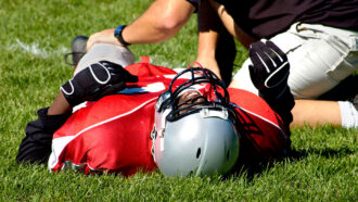 a teen football player wearing full protective gear lays on his back on a field, face scrunched up in pain, as someone kneels next to him