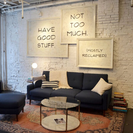 A living room. A dark blue sofa and coffee table on an oriental rug in front of a white-painted brick wall. On the wall three signs read 'Have good stuff', 'Not too much', '(Mostly reclaimed)'