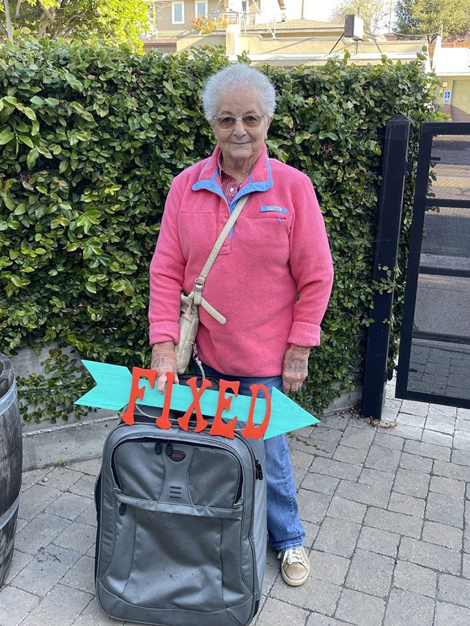 A senior woman stands outside with a roller bag. She is smiling and propping up a 'Fixed' sign on the bag.