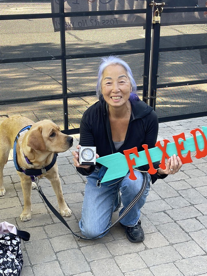 a happy older Asian woman with white hair and purple highlights shows off an electronic device while holding a 'FIXED' sign. Her dog is standing next to her with a quizical look. 