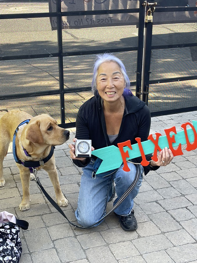 a happy older Asian woman with white hair and purple highlights shows off an electronic device while holding a 'FIXED' sign. Her dog is standing next to her with a quizical look.