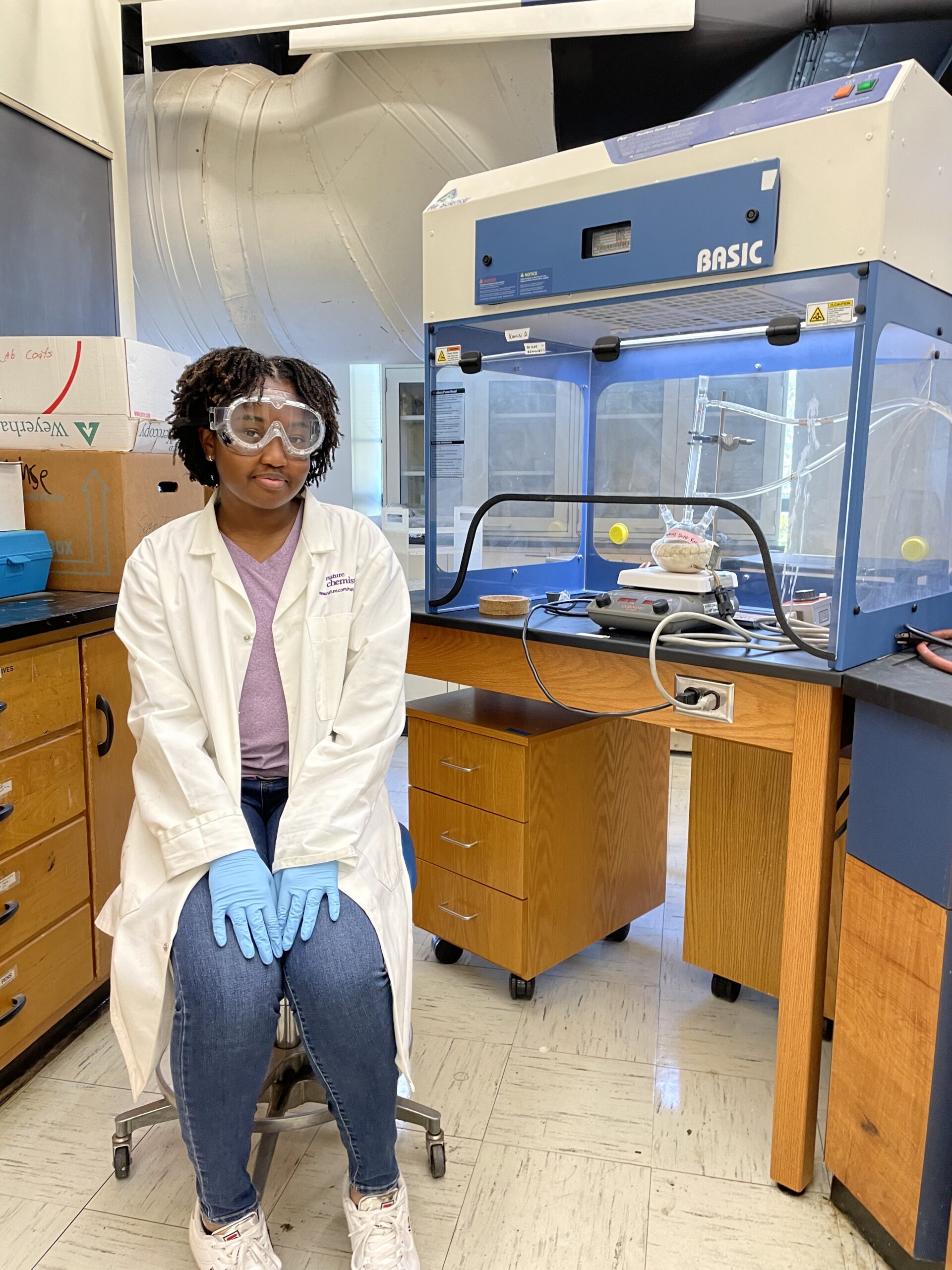 a teen girl wearing a lab coat, goggles and rubber gloves sits next to a desk full of complex machinery