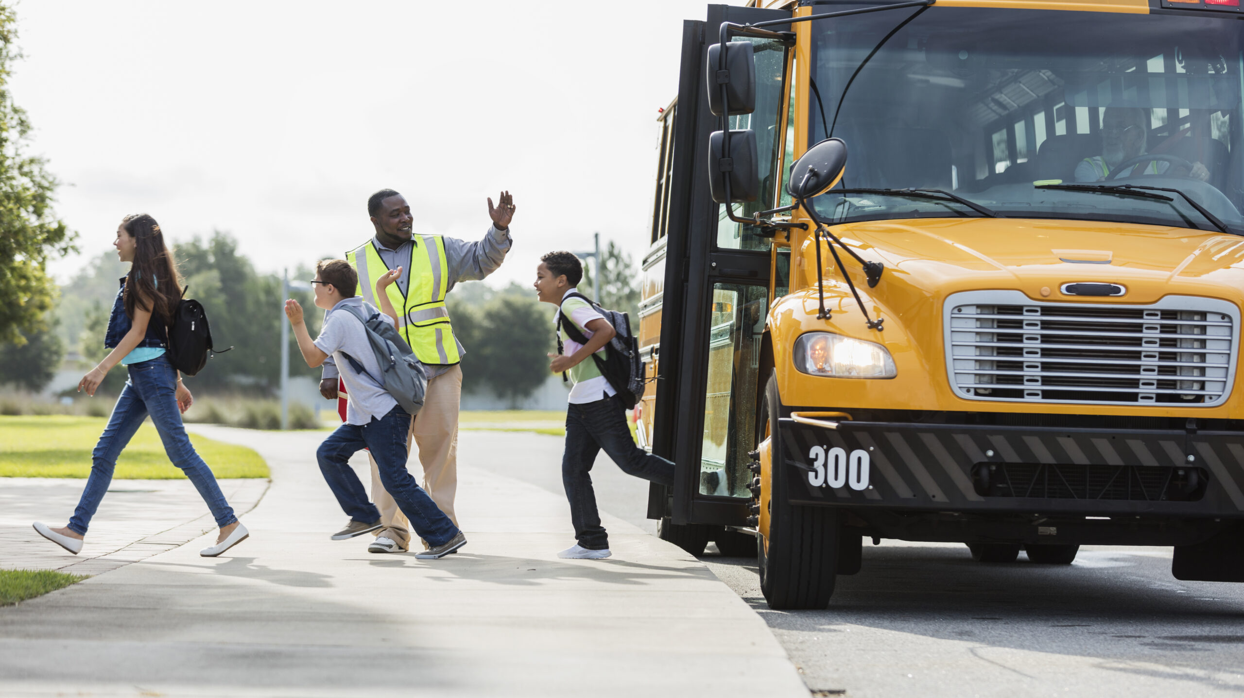 A group of three middle school students, 12 and 13 years old, exiting a yellow school bus. The crossing guard is waving to the bus driver sitting inside the bus. 