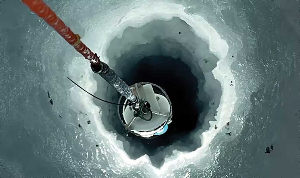 A photo of a research instrument being lowered down by an orange cord in to the ice through a borehole.
