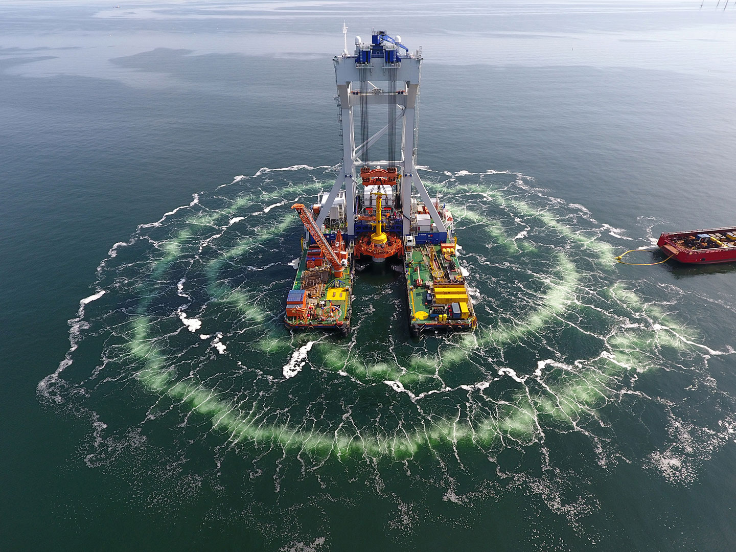 an aerial view of an offshore windfarm being constructed in the ocean. Around the platform being built are rings of bubbles expanding outward into the ocean.