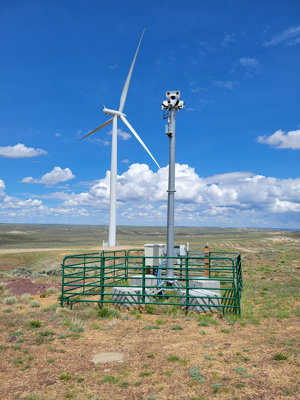 A wind turbine is in the distance, the landscape is flat and scrubby. In the foreground a green metal fence surrounds a small tower equipped with cameras.