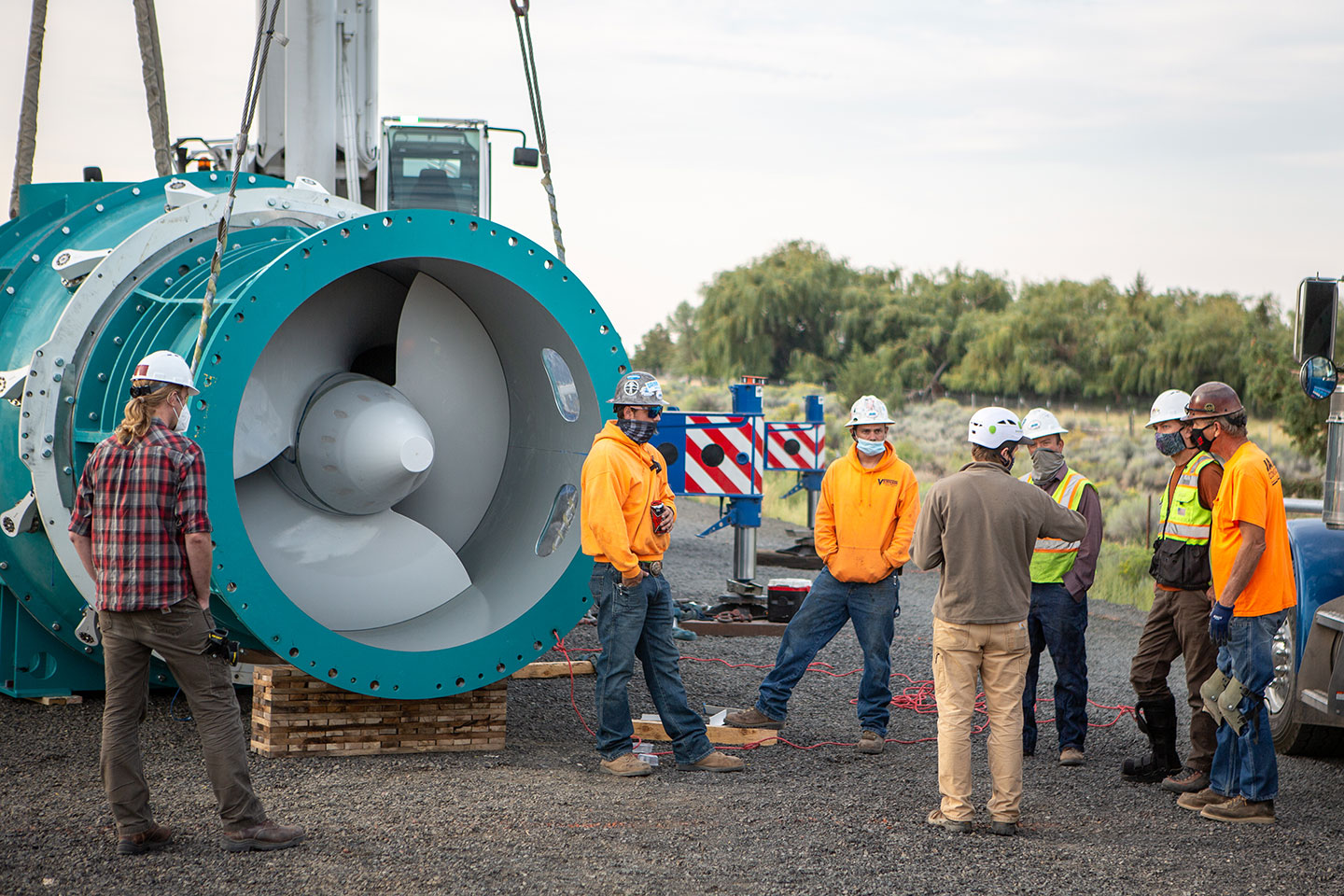 A photo of turbine to be installed at a Hydro Plant. The turbine is taller than a person and has curved blades. It's housed inside a metal cylinder and almost looks like a jet engine. There are a lot of people standing around nearby wearing construction safety gear.