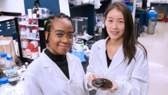 A photo of two women scientists in a lab smiling at the camera. Néhémie Guillomaitre, on the left, is a Black woman with large round wireframe glasses and her hair braided away from her face. Xiaohui Xu, an Asian woman with shoulder length dark hair, stands on the rigth and is holding a petri dish full of the new hydrogel.