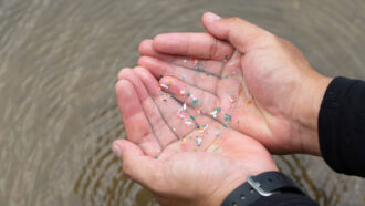 a pair of cupped palms hold a bit of water scooped out of a river that contains multicolored bits of plastic