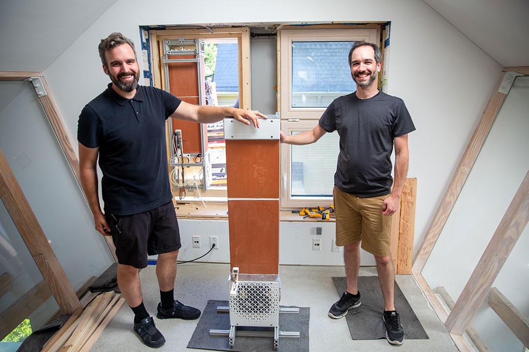 A photo of two men standing on either side of a narrow electronic tower (the evaporative AC). Both are smiling at the camera.