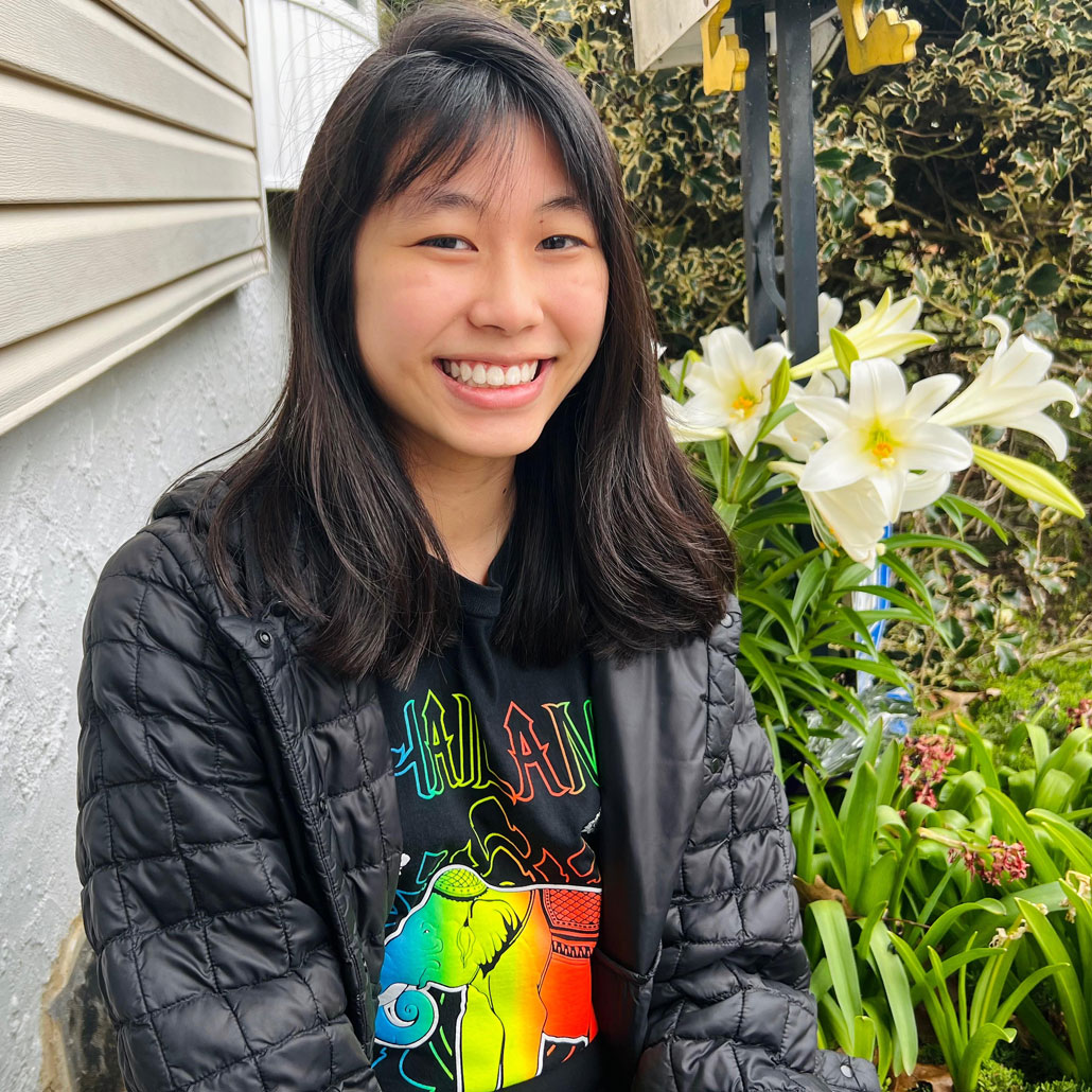 a photo of Natasha Kulviwat, an Asian teen with shoulder length black hair. She is wearing a black puffer jacket, a t-shirt that says "Thailand" on it in rainbow print, and she is smiling at the camera. She's standing outside next to some lillies.