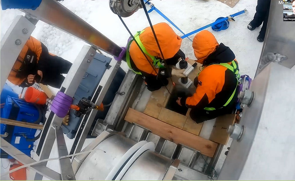 An overhead photo showing scientists in orange parkas lowering instruments into a borehole leading to the ice cavern. 