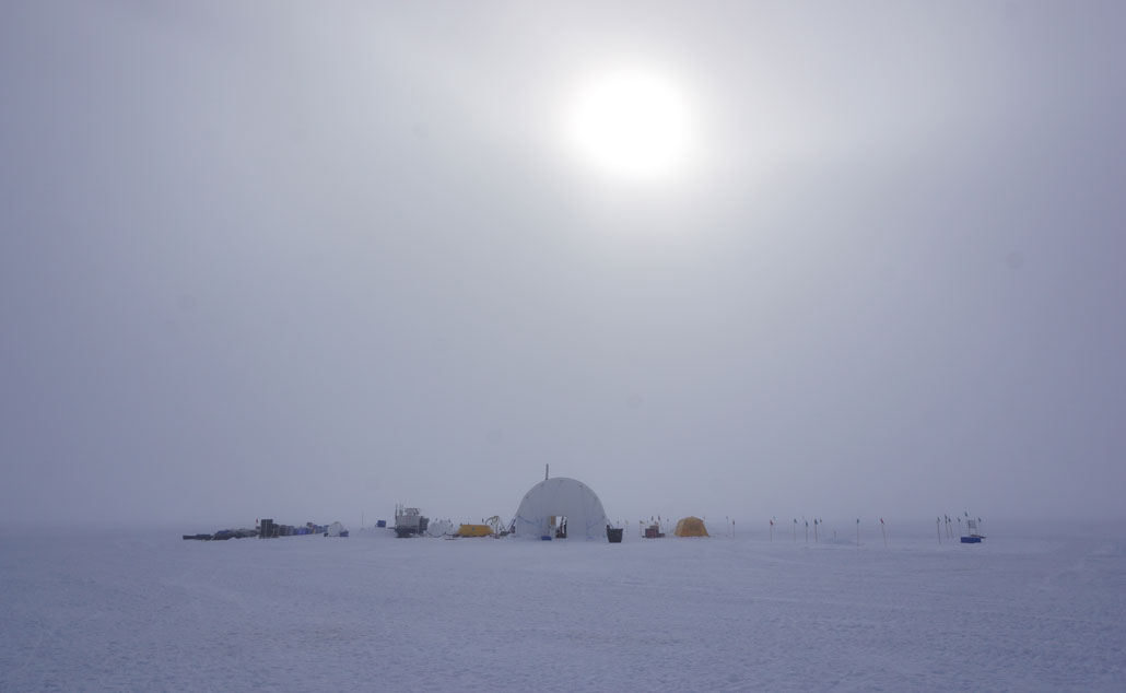 A photo of a domed tent marking a camp on the Kamb Ice Stream. Various scientific equipment and containers are scattered around the tent on the snow and ice. The sky is overcast, the sun is shining through thick cloud cover and the ground is white. 