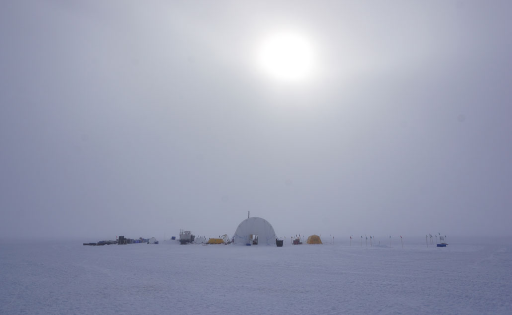A photo of a domed tent marking a camp on the Kamb Ice Stream. Various scientific equipment and containers are scattered around the tent on the snow and ice. The sky is overcast, the sun is shining through thick cloud cover and the ground is white.