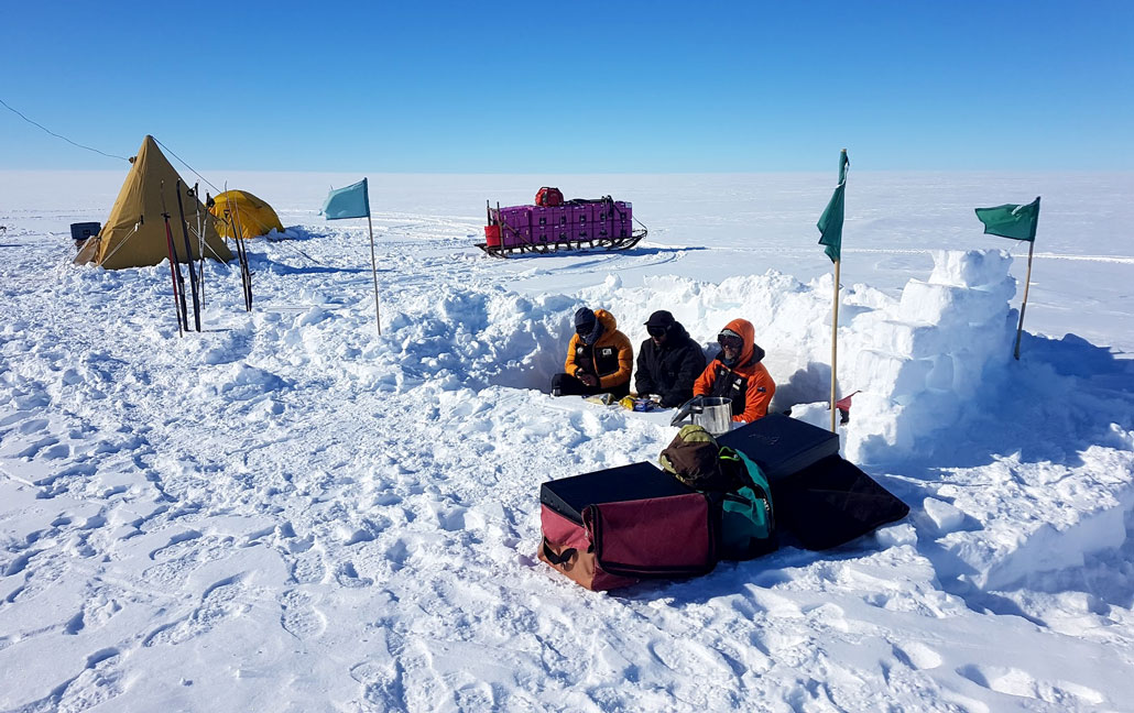 A photo of three scientists bundled up in cold weather gear, they are sitting down and eating while sheltered in a pit behind a snow wall. The wall is made of large snow bricks and curves slightly around the pit. Green flags on stakes mark the outer edges of the snow wall. Tents are behind the wall and to the left.