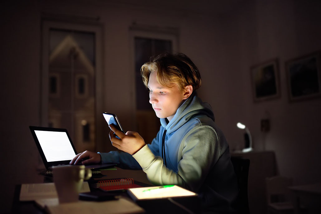 a young man up late at night, he is sitting in a dark room and working on his laptop, while also using his phone