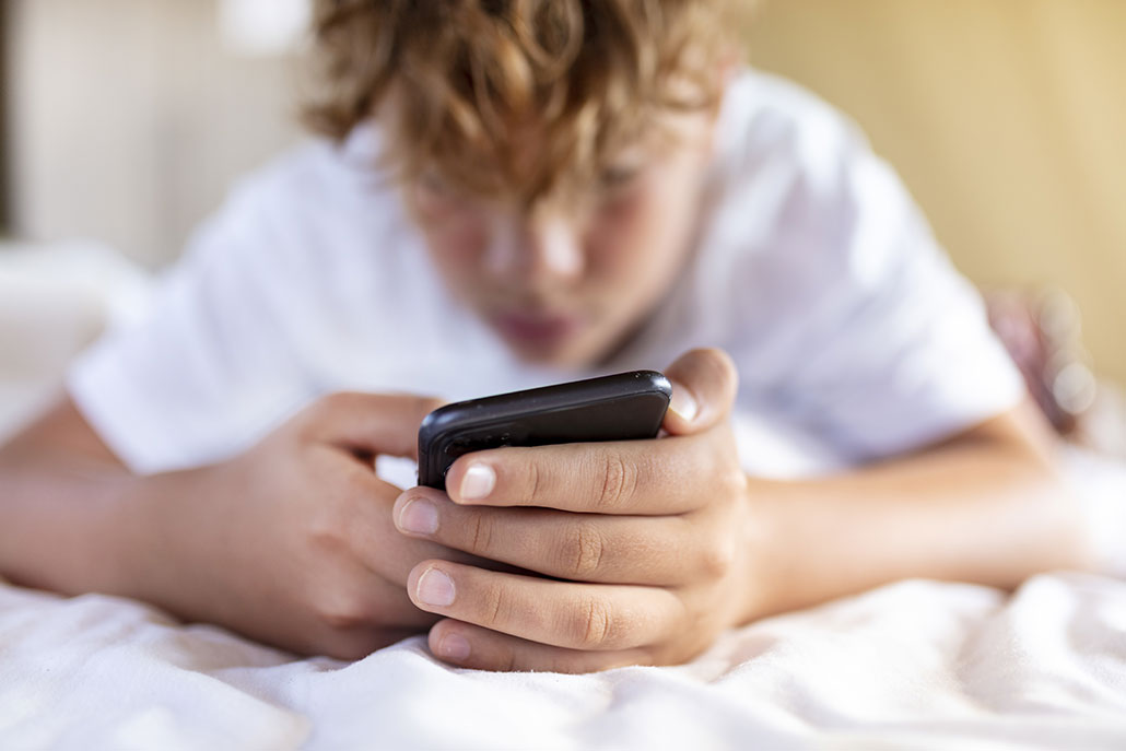 a photo of a young man with blond hair lying on a bed and using his smart phone. He's wearing a white shirt and lying on his stomach on white sheets. The only part of the image in focus is his hands holding the cell phone.