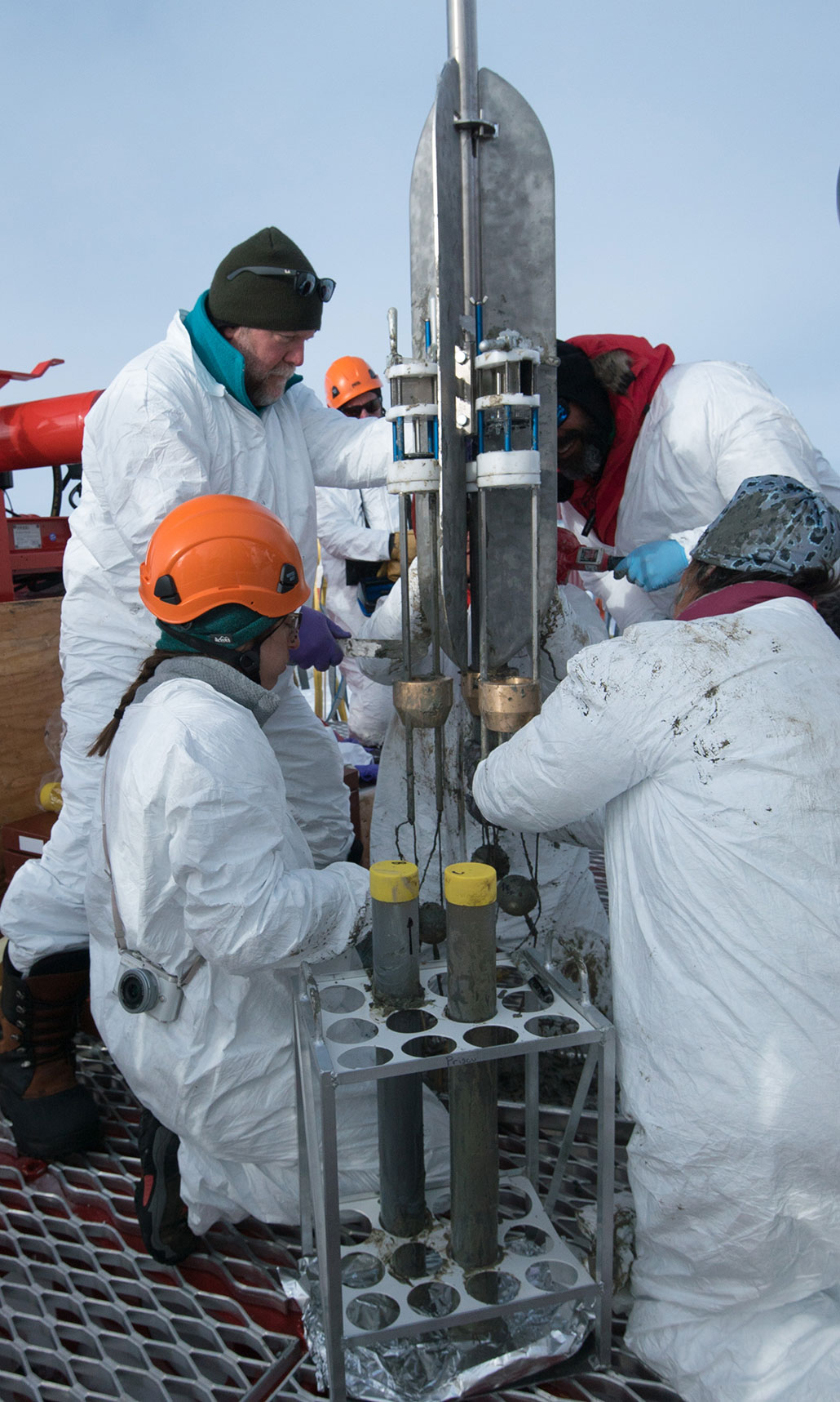 A photo of four researchers around the apparatus used to bring sediment cores back up to the surface of Lake Mercer. Two sediment cores in clear tubes are in a stand next to the apparatus.