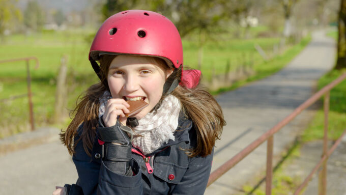 a middle-school girl with brown hair, a red helmet and a jacket bites into a chocolate bar while standing outside, leaning against a fence