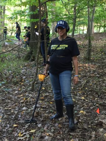 A pregnant Kelly Knight stands in the middle of a forest. She is holding a metal detector and hearing headphones